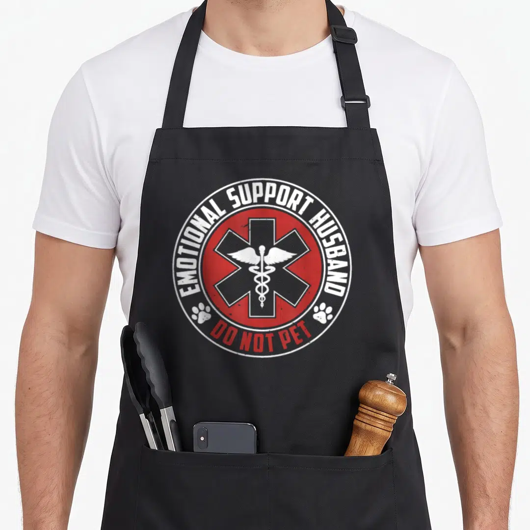 Emotional couple apron - Red circle with Star of Life, caduceus, and text EMOTIONAL SUPPORT HUSBAND DO NOT PET — shown on a black cotton canvas apron against a white background