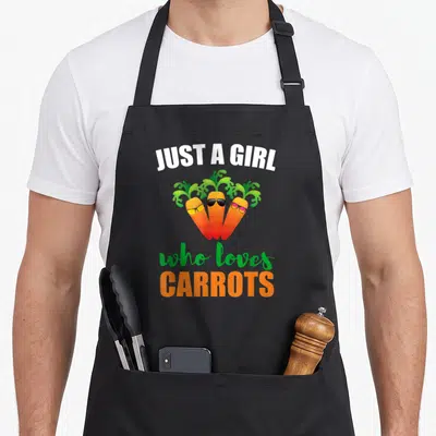 Funny carrot vegetable apron - Three orange carrots wearing sunglasses under text JUST A GIRL who loves CARROTS — shown on a black cotton canvas apron against a white background