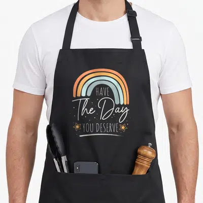 Rainbow boho apron - Four color rainbow arch above stacked text HAVE The Day YOU DESERVE flanked by two stars — shown on a black cotton canvas apron against a white background