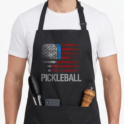 Flag pickleball apron - Pickleball paddle shaped like a distressed US flag with red white stripes and blue star field — shown on a black cotton canvas apron against a white background