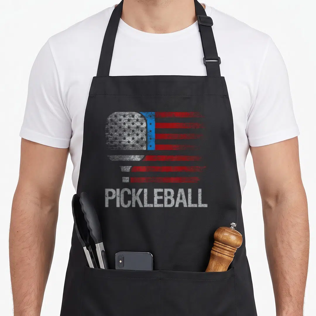 Flag pickleball apron - Pickleball paddle shaped like a distressed US flag with red white stripes and blue star field — shown on a black cotton canvas apron against a white background
