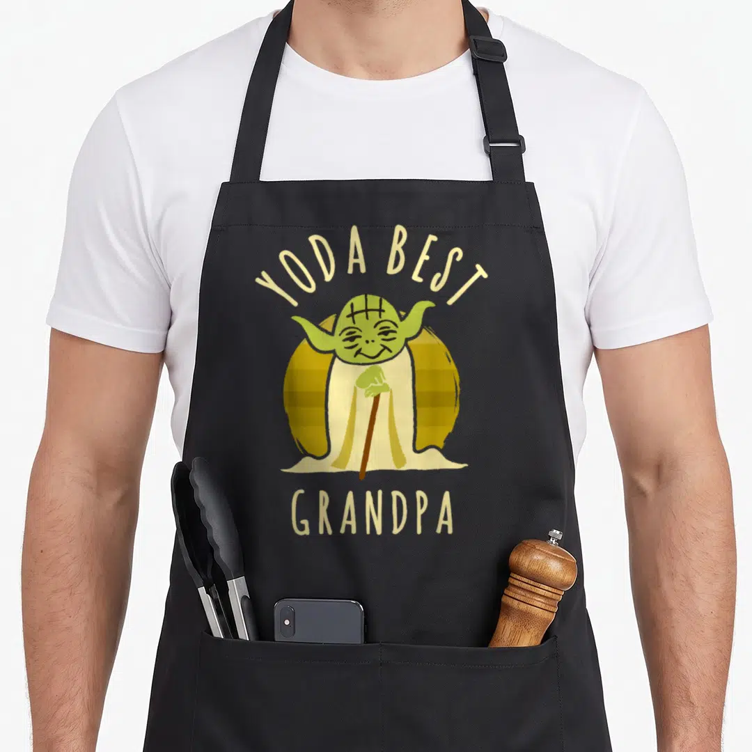 Grandpa cartoon apron - Yoda holding cane with YODA BEST GRANDPA text arched above and below — shown on a black cotton canvas apron against a white background