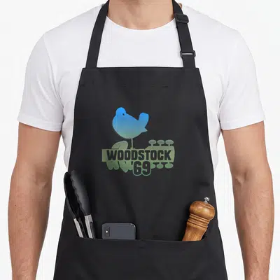 Woodstock love dove apron - Blue and green bird silhouette above stacked WOODSTOCK 69 text — shown on a black cotton canvas apron against a white background