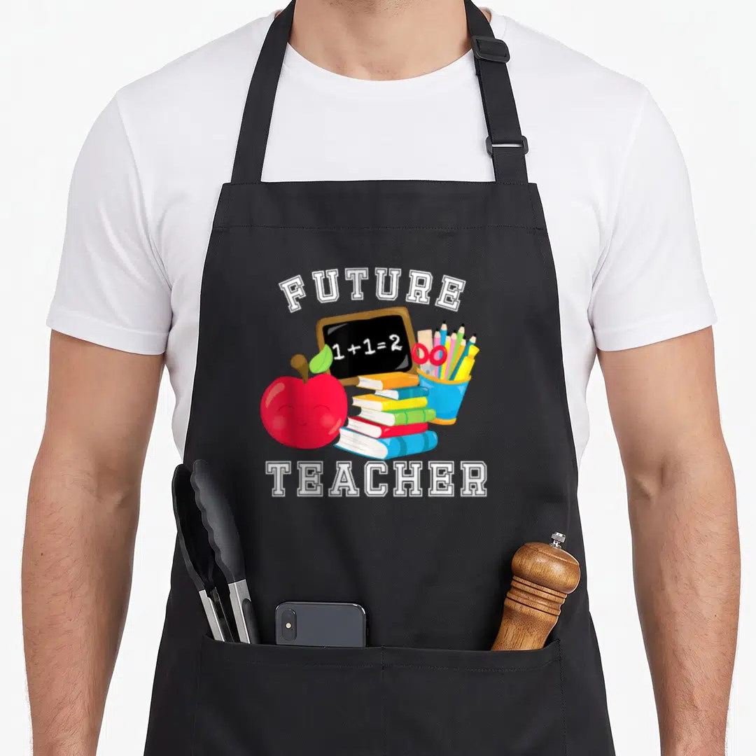 Future costume teacher apron - Cartoon red apple, chalkboard with 1+1=2, stacked books, and pencil cup framed by white — shown on a black cotton canvas apron against a white background