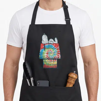 Cute dye tie apron - Snoopy lying on a red blue yellow and green tie-dye doghouse — shown on a black cotton canvas apron against a white background