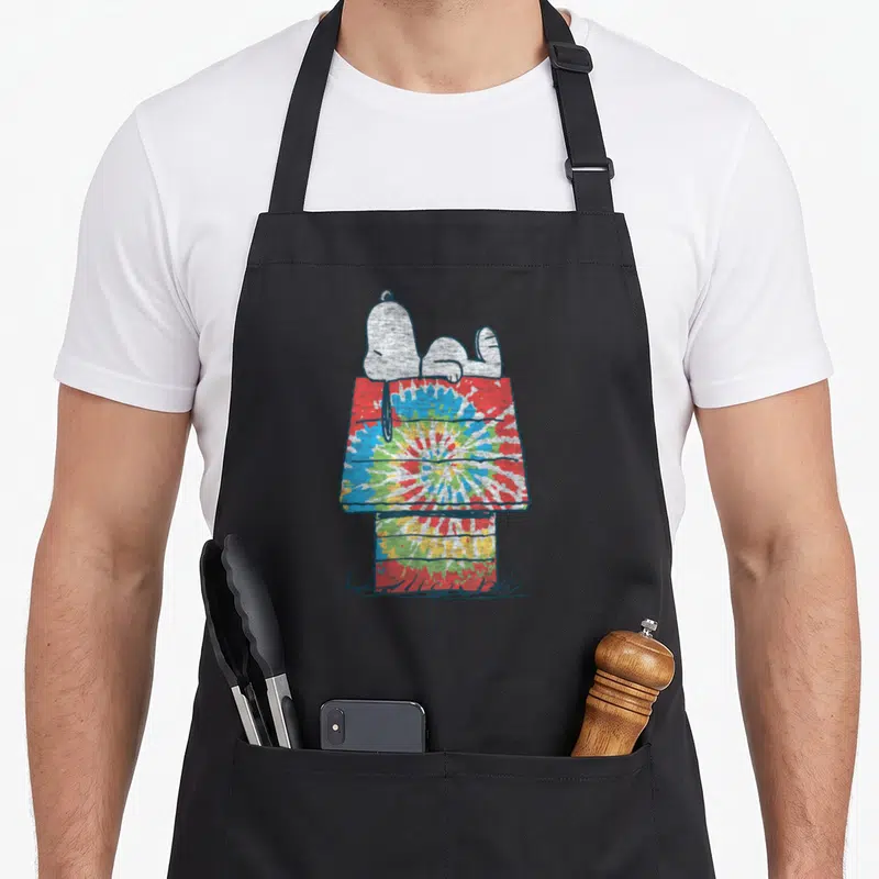 Cute dye tie apron - Snoopy lying on a red blue yellow and green tie-dye doghouse — shown on a black cotton canvas apron against a white background