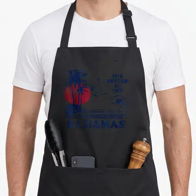 Better 70s apron - Distressed blue text It's Better in the Bahamas with two birds, palm tree, red sun, and wave — shown on a black cotton canvas apron against a white background