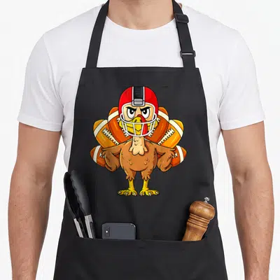 Player thanksgiving apron - Cartoon turkey in red helmet with footballs resembling tail feathers — shown on a black cotton canvas apron against a white background