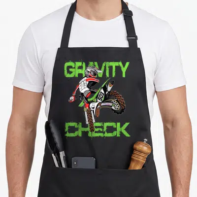 Dirt rider motocross bike apron - Cartoon motocross rider doing a wheelie framed by green Gravity Check text — shown on a black cotton canvas apron against a white background