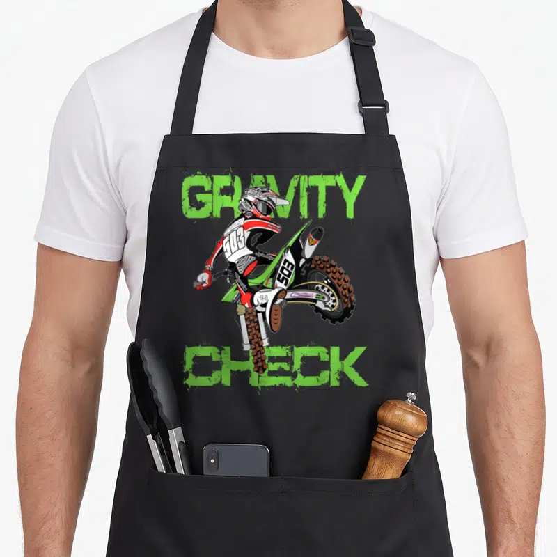 Dirt rider motocross bike apron - Cartoon motocross rider doing a wheelie framed by green Gravity Check text — shown on a black cotton canvas apron against a white background
