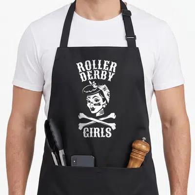 Roller pinup apron - White ROLLER DERBY GIRLS text above a tattooed woman with crossed bones — shown on a black cotton canvas apron against a white background