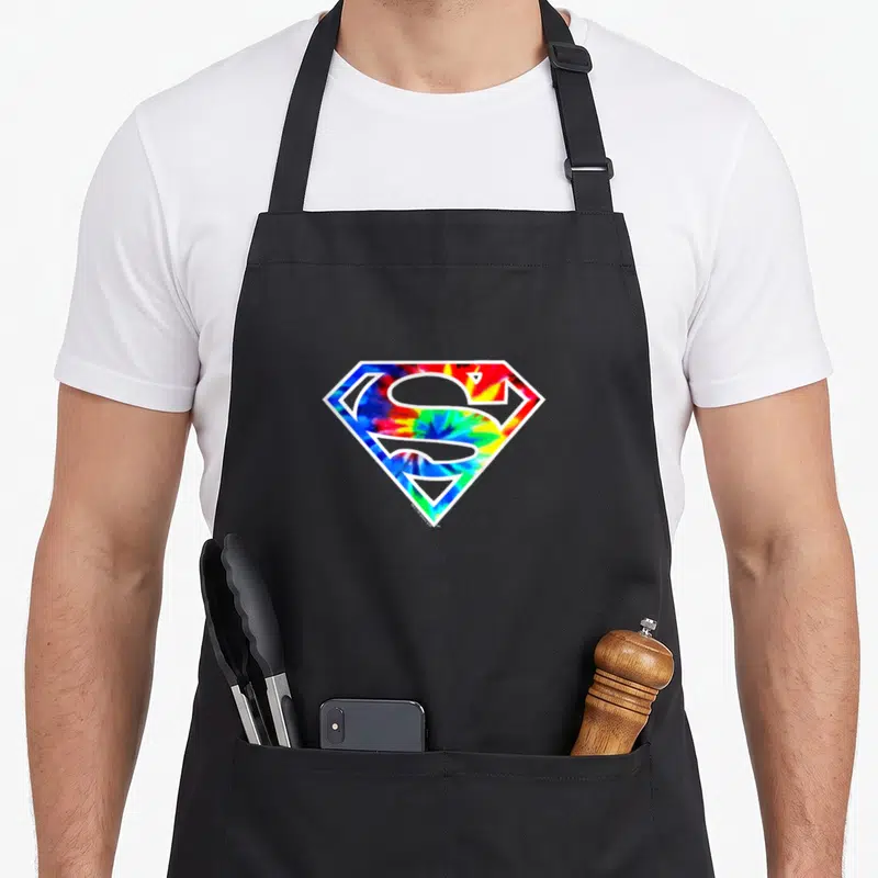 Vintage dye tie apron - Superman shield logo with blue red yellow and green tie-dye fill — shown on a black cotton canvas apron against a white background