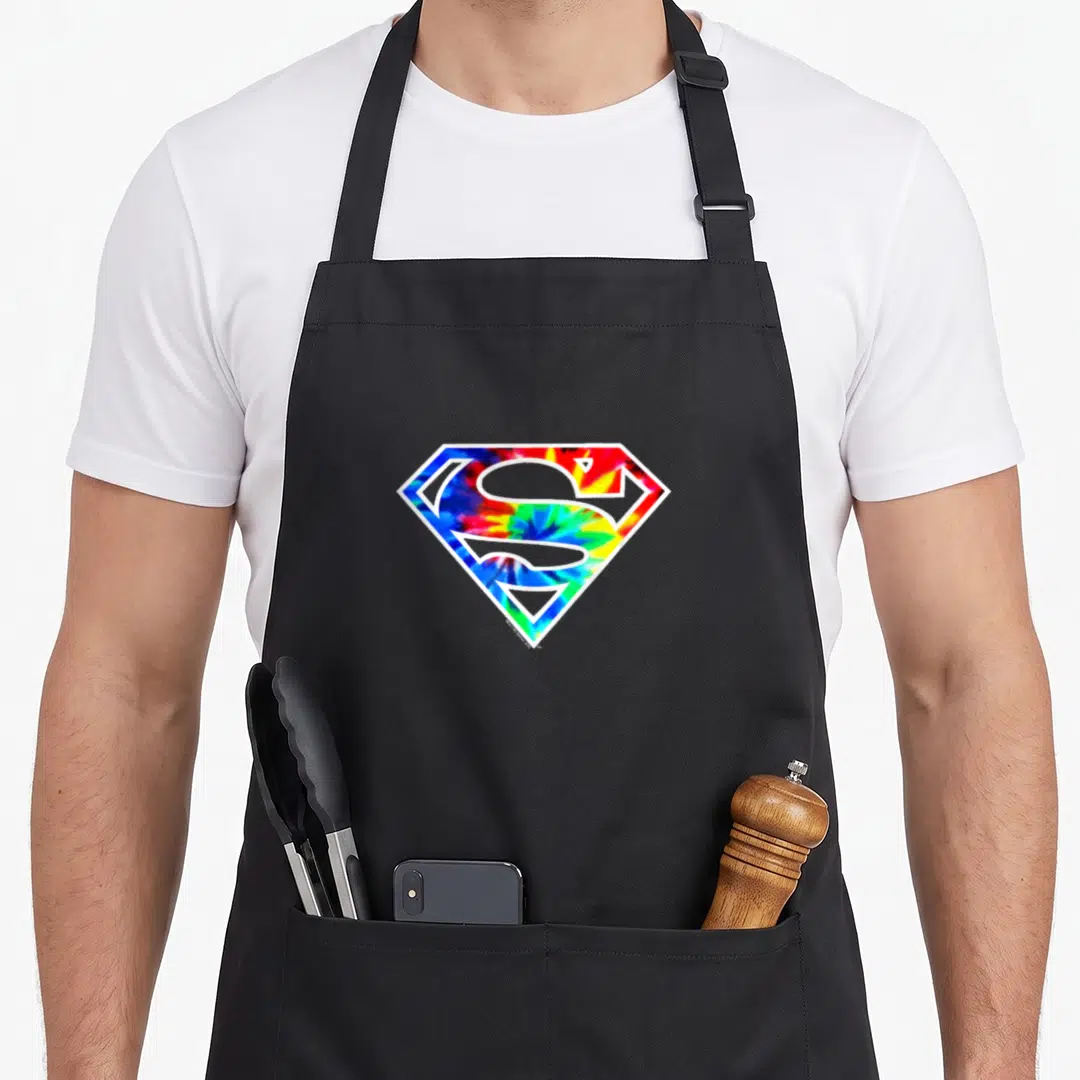 Vintage dye tie apron - Superman shield logo with blue red yellow and green tie-dye fill — shown on a black cotton canvas apron against a white background