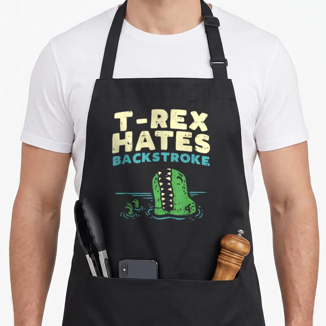 Backstroke dinosaur apron - T-Rex Hates Backstroke text above green dinosaur head and small arms in teal water — shown on a black cotton canvas apron against a white background