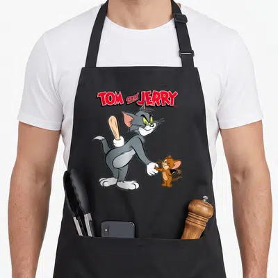 Hand crew neck apron - Tom and Jerry cartoon characters shaking hands, cat hiding a bat, mouse holding a bomb — shown on a black cotton canvas apron against a white background
