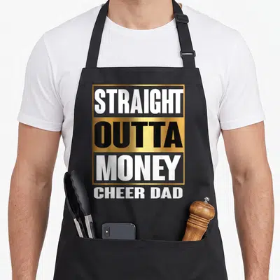 Straight outta money apron - Stacked gold and white text reading Straight Outta Money Cheer Dad on black background — shown on a black cotton canvas apron against a white background