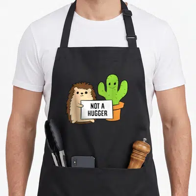 Hedgehog cactus apron - Cartoon hedgehog and cactus holding white sign reading NOT A HUGGER — shown on a black cotton canvas apron against a white background