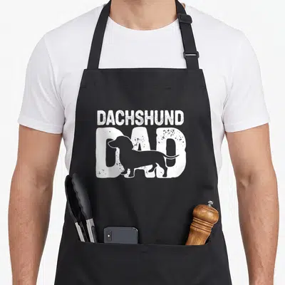 Doxie father dog apron - White distressed DACHSHUND DAD text with dachshund silhouette on black bib — shown on a black cotton canvas apron against a white background