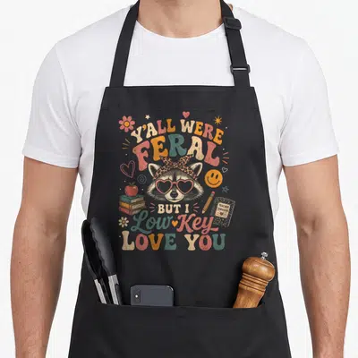 Yall coffee apron - Raccoon in heart sunglasses surrounded by books and text Y'all Were Feral But I Love You — shown on a black cotton canvas apron against a white background