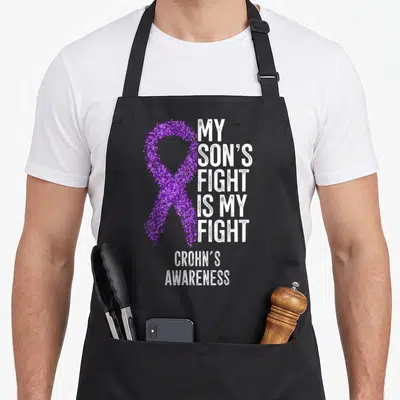 Battle crohns awareness apron - Purple dotted ribbon and white text MY SON'S FIGHT IS MY FIGHT CROHN'S AWARENESS — shown on a black cotton canvas apron against a white background