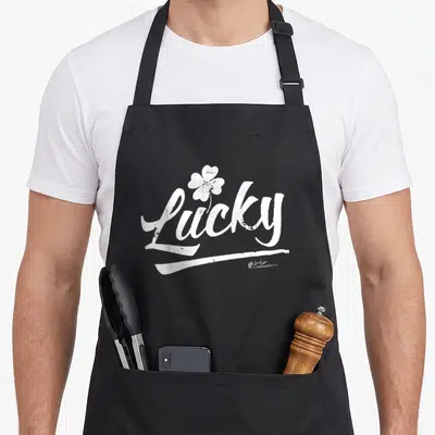 Cute lucky apron - White distressed script Lucky with four-leaf clover and brushstroke underline — shown on a black cotton canvas apron against a white background