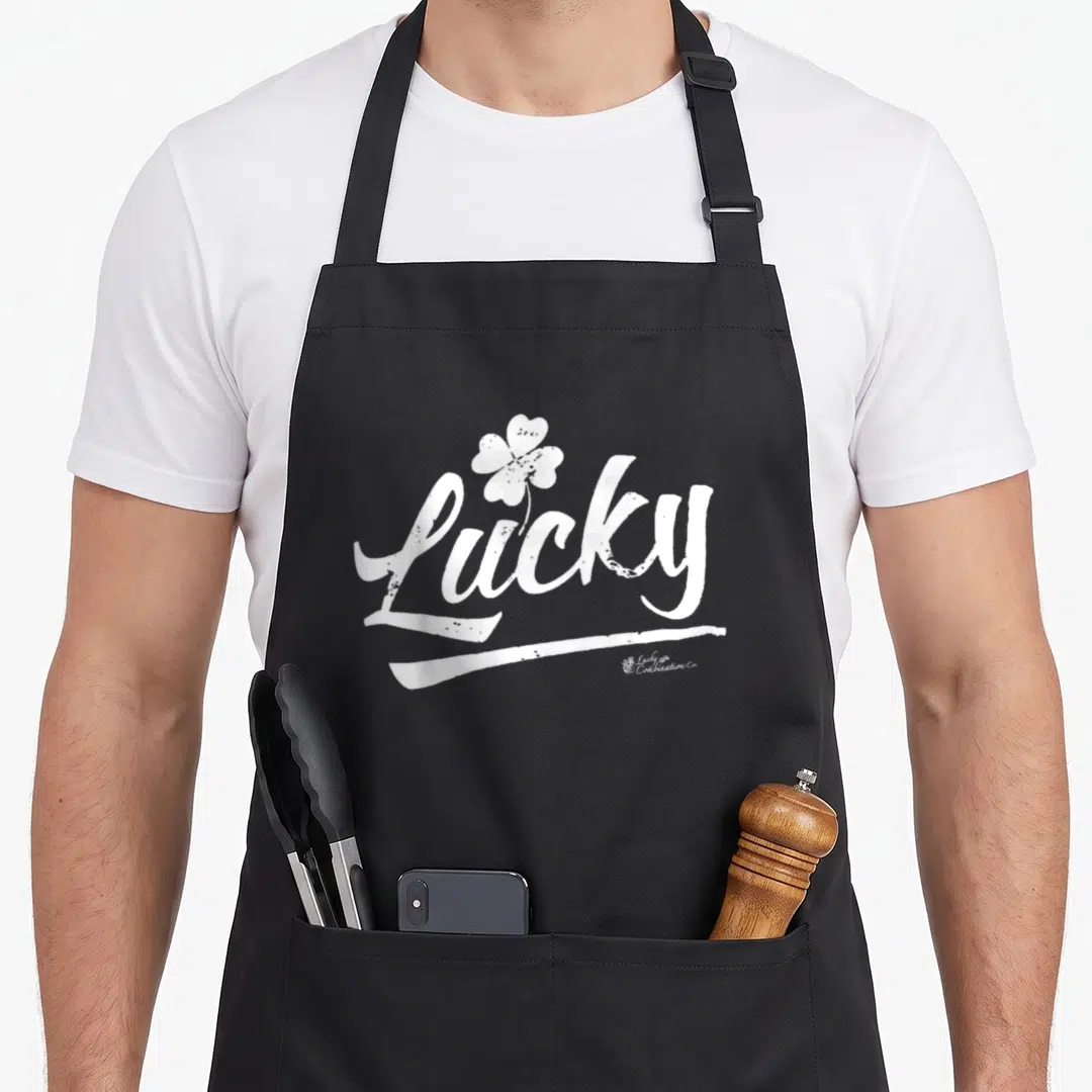 Cute lucky apron - White distressed script Lucky with four-leaf clover and brushstroke underline — shown on a black cotton canvas apron against a white background