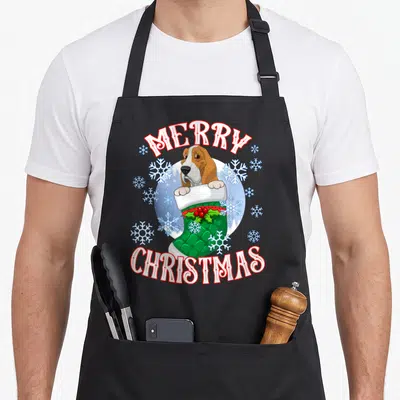Basset womens christmas aprons - Cartoon basset hound in green stocking with Merry Christmas text and snowflakes — shown on a black cotton canvas apron against a white background