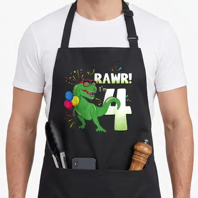 Birthday dinosaur t-rex apron - Green dinosaur with red sunglasses holding balloons next to number 4 and RAWR text — shown on a black cotton canvas apron against a white background