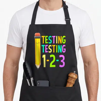 Testing teacher apron - Cartoon pencil next to stacked yellow green purple blue pink text TESTING TESTING 1-2-3 — shown on a black cotton canvas apron against a white background