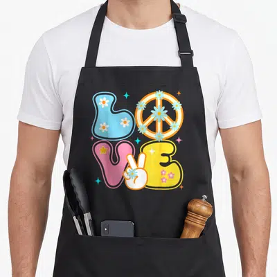 Sign 60s tie-dye apron - LOVE text with peace sign O, hand holding V, and scattered daisies in blue pink yellow orange — shown on a black cotton canvas apron against a white background