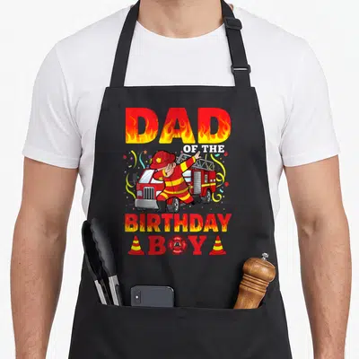 Truck firefighter aprons - DAD OF THE BIRTHDAY BOY text with dabbing firefighter and fire truck — shown on a black cotton canvas apron against a white background