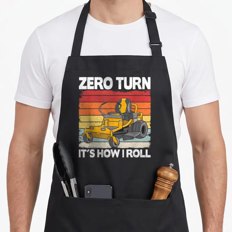 Zero hawk dad apron - Retro yellow zero turn mower on red orange striped background with white text — shown on a black cotton canvas apron against a white background