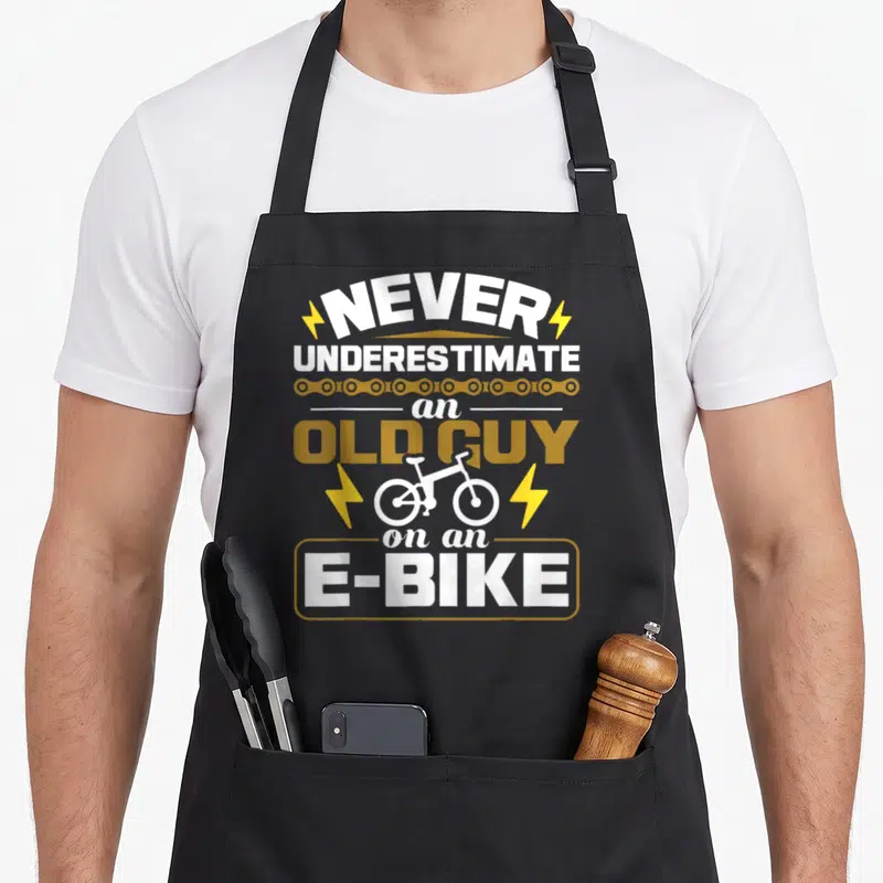 Underestimate bicycle apron - Stacked text Never Underestimate an Old Guy On an E-Bike with e-bike icon and lightning — shown on a black cotton canvas apron against a white background