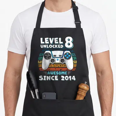 Eight dad and son aprons - White game controller over pixelated rainbow arc with Level 8 Unlocked text — shown on a black cotton canvas apron against a white background