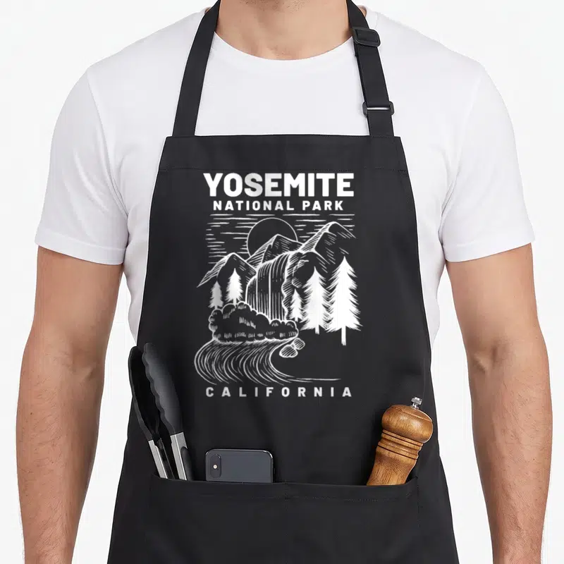California national park apron - White line art of Yosemite mountains waterfall and river with stacked text on black — shown on a black cotton canvas apron against a white background