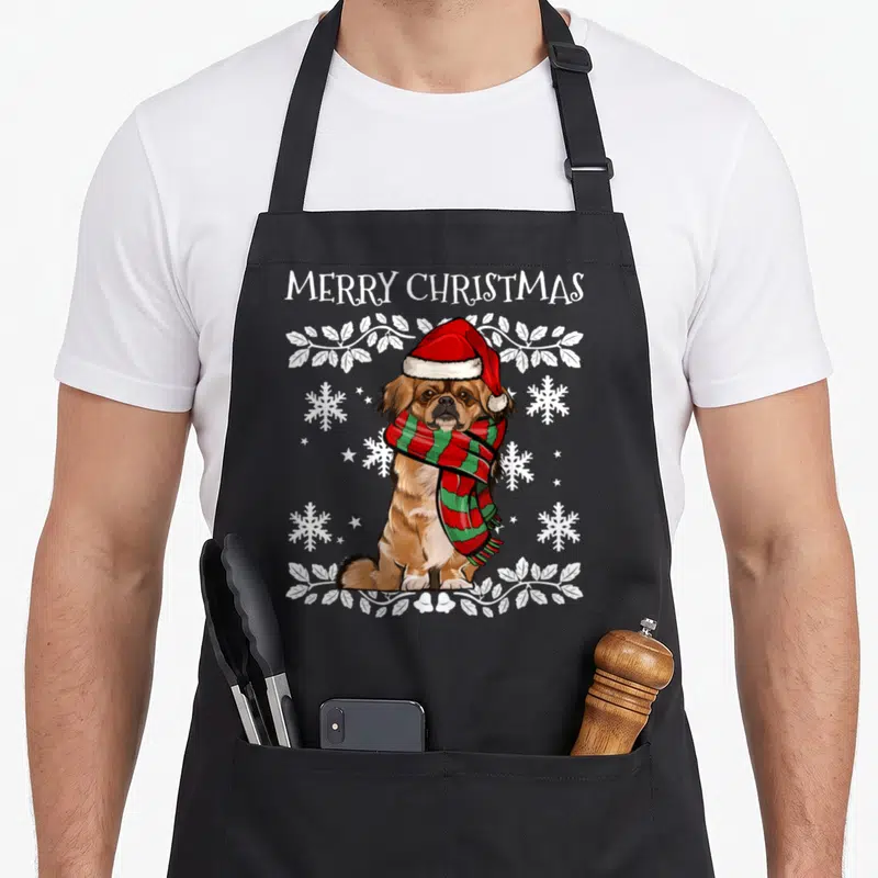 Ornament merry christmas apron - Realistic brown Pekingese dog wearing red hat and green scarf amidst white snowflakes — shown on a black cotton canvas apron against a white background