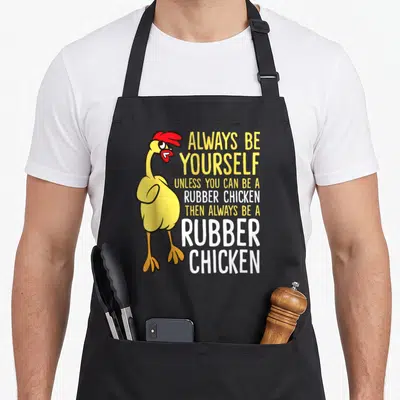 Alway chicken apron - Cartoon yellow rubber chicken with red details next to stacked yellow and white text — shown on a black cotton canvas apron against a white background