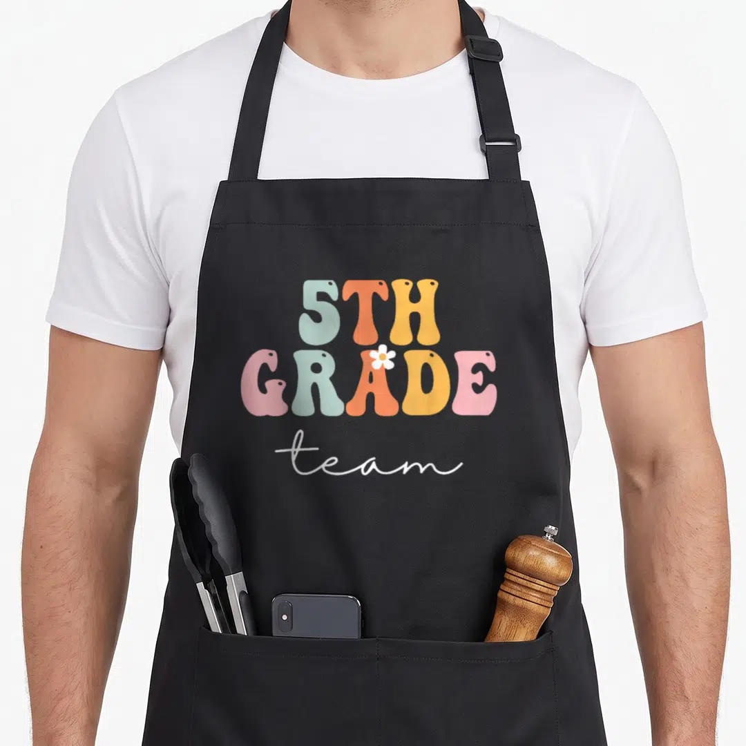 5th groovy grade apron - Retro 5TH GRADE team text in blue orange yellow pink and white with a flower — shown on a black cotton canvas apron against a white background