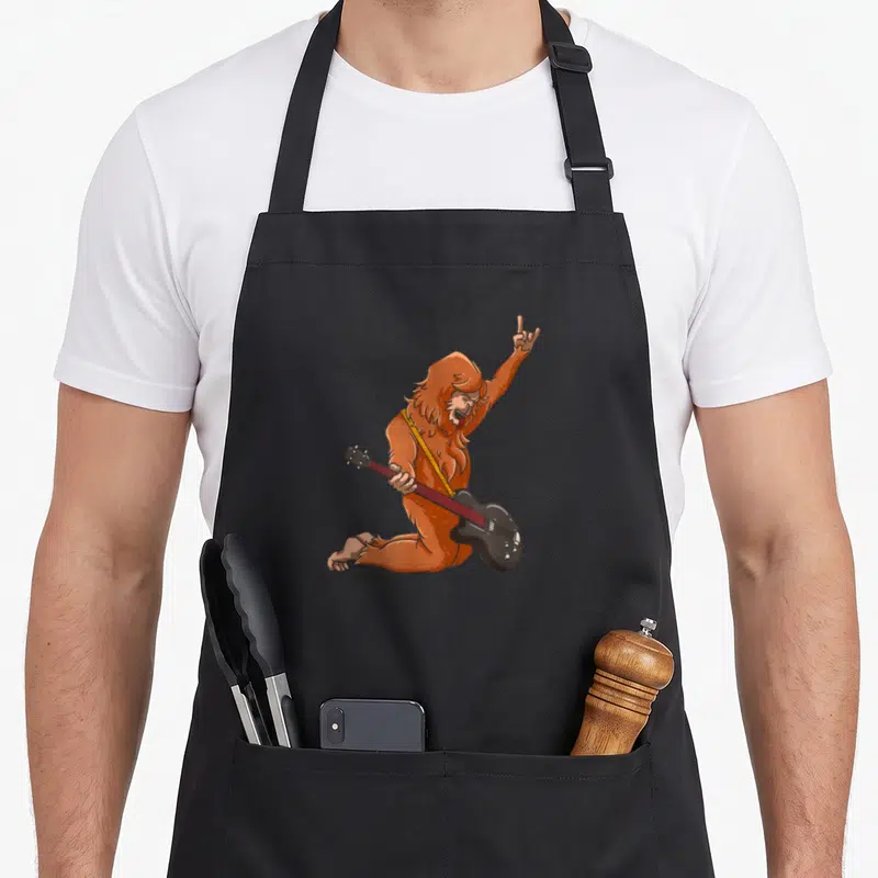 Bassist bigfoot apron - Cartoon brown bigfoot kneeling playing black guitar with yellow strap and raised rock hand — shown on a black cotton canvas apron against a white background