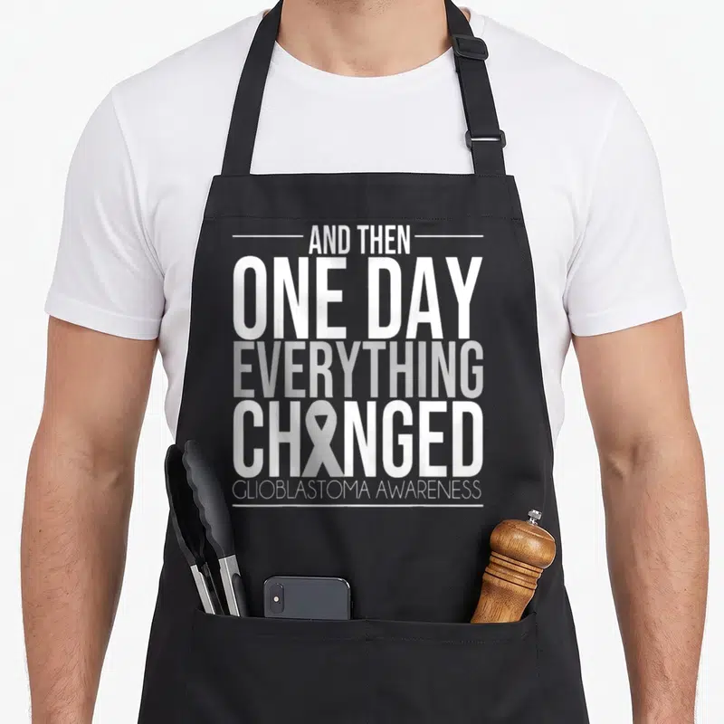 Glioblastoma brain apron - Stacked white and grey text reading AND THEN ONE DAY EVERYTHING CHANGED GLIOBLASTOMA — shown on a black cotton canvas apron against a white background
