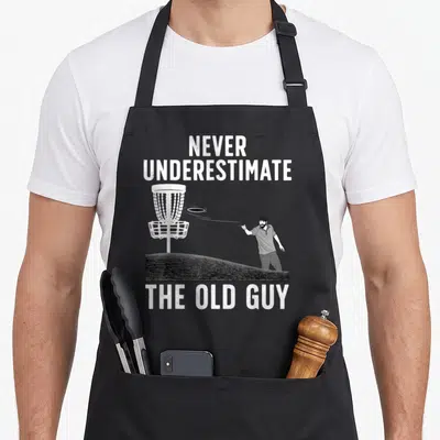 Grandpa golf apron - White NEVER UNDERESTIMATE THE OLD GUY text above a man throwing a disc at a basket on a grassy hill — shown on a black cotton canvas apron against a white background