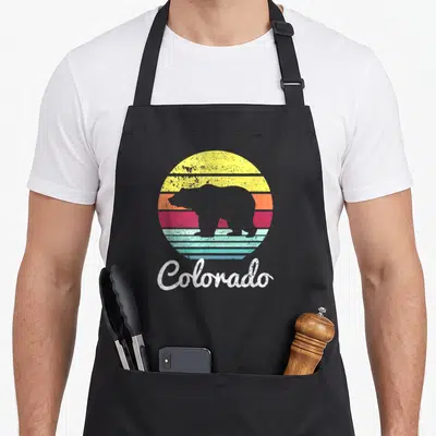 Wildlife colorado apron - Retro bear silhouette over yellow orange red teal striped circle with Colorado text below — shown on a black cotton canvas apron against a white background