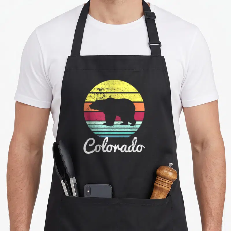 Wildlife colorado apron - Retro bear silhouette over yellow orange red teal striped circle with Colorado text below — shown on a black cotton canvas apron against a white background