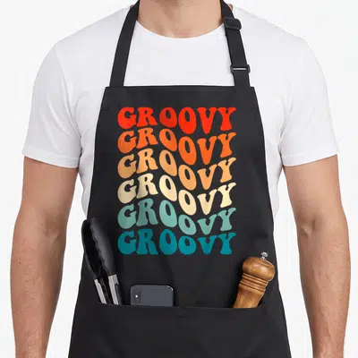 Groovy hippie apron - Six stacked lines of GROOVY text in red orange cream and blue typography — shown on a black cotton canvas apron against a white background
