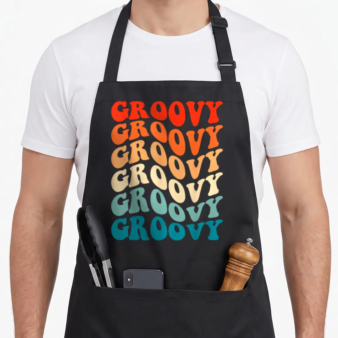 Groovy hippie apron - Six stacked lines of GROOVY text in red orange cream and blue typography — shown on a black cotton canvas apron against a white background