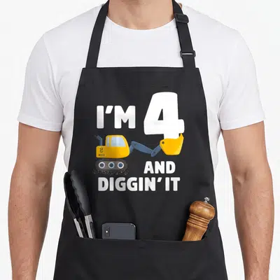 Birthday four truck apron - I'M 4 AND DIGGIN' IT text with yellow excavator and blue details — shown on a black cotton canvas apron against a white background