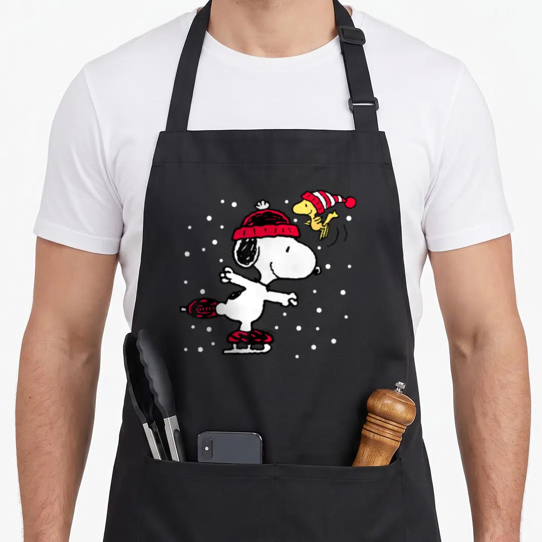 Skate holiday apron - Snoopy ice skating in red hat with Woodstock and white snowflakes on black fabric — shown on a black cotton canvas apron against a white background