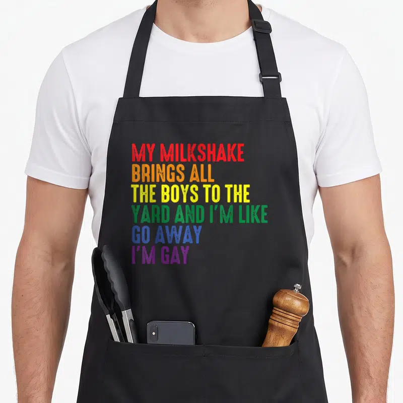 Milkshake gay apron - Rainbow stacked text reading My Milkshake Brings All The Boys To The Yard I'm Gay — shown on a black cotton canvas apron against a white background