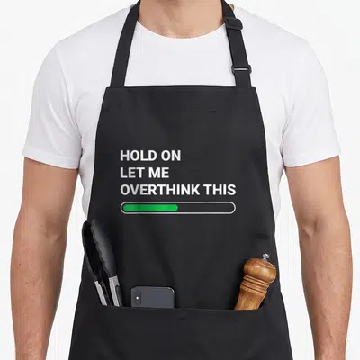 Overthink sarcastic apron - White text reading Hold On Let Me Overthink This above a green loading bar — shown on a black cotton canvas apron against a white background