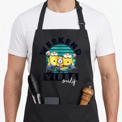 Despicable weekend vibes apron - Three yellow minions holding ice cream and a party horn under arched text over a teal — shown on a black cotton canvas apron against a white background